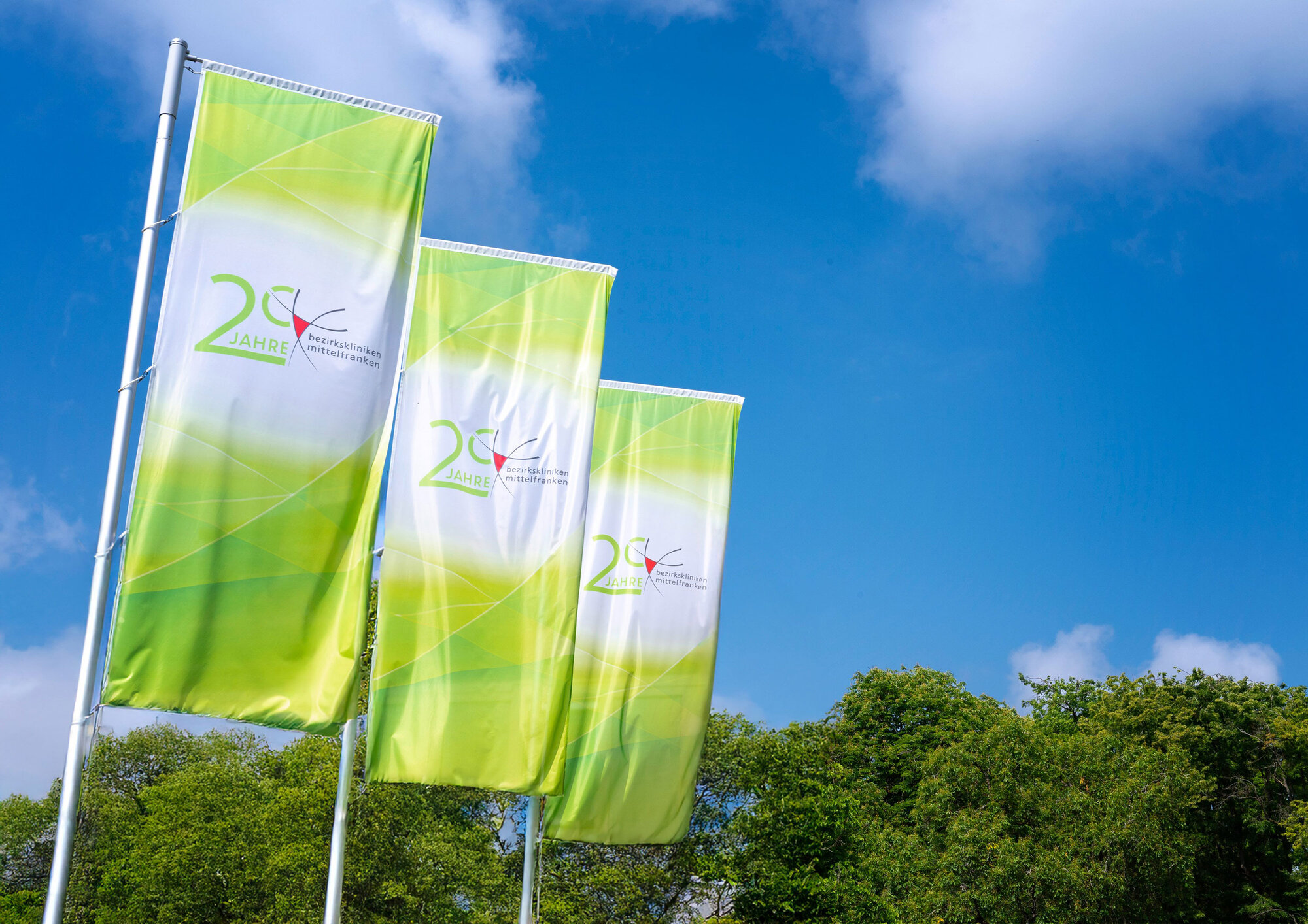 Drei grüne Fahnen mit Jubiläumslogo der Bezirkskliniken Mittelfranken wehen im Wind.