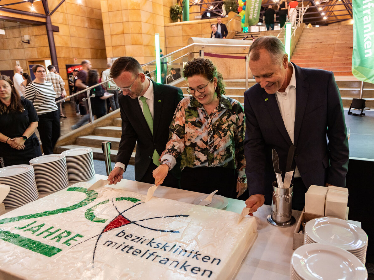 Peter Daniel Forster, Bezirkstagspräsident und Vorsitzender des Verwaltungsrats, Kaufmännische Vorständin Stinne Fronius und Dr. Matthias Keilen, Vorstand Medizin schneiden eine riesige Torte zum 20. Geburtstag der Bezirkskliniken an