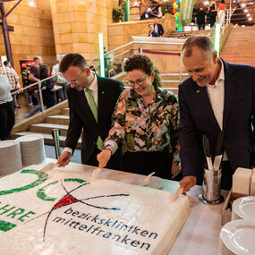 Peter Daniel Forster, Bezirkstagspräsident und Vorsitzender des Verwaltungsrats, Kaufmännische Vorständin Stinne Fronius und Dr. Matthias Keilen, Vorstand Medizin schneiden eine riesige Torte zum 20. Geburtstag der Bezirkskliniken an